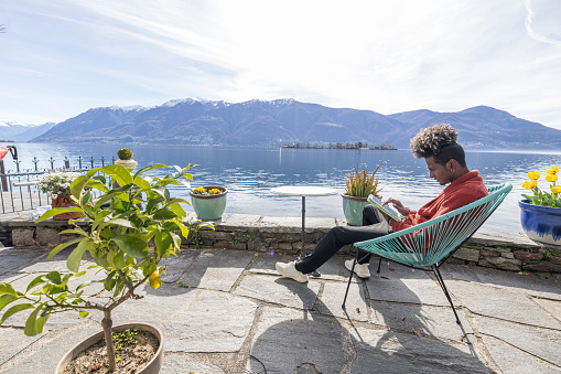 Side view of a young man relaxed on a patio chair, working on his tablet outdoors. He has a beautiful view of a mountain, a lake and is surrounded by potted plants.