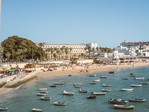 Boats anchored and people sunbathing on a sandy summer beach Playa de La Caleta in Cadiz, Andalusia, Spain