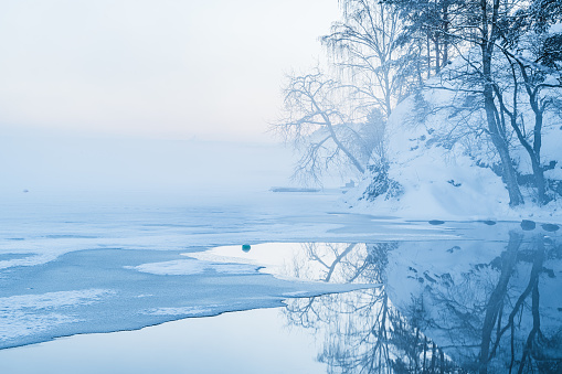 A body of water in Mölndal, Sweden is surrounded by snow-covered trees. The trees cast reflections on the calm Lake Rådasjön, creating a serene winter scene in the misty atmosphere.