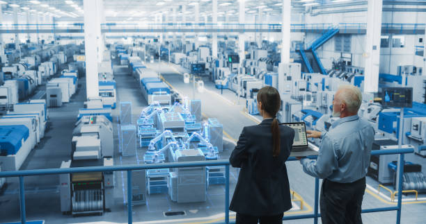 istock Male and Female Standing at Electronics Factory, Using Computer and Having a Conversation. Augmented Reality Visualization of a Conveyor Belt Production Line with Robot Arms Appears In Front of Them 2143504970