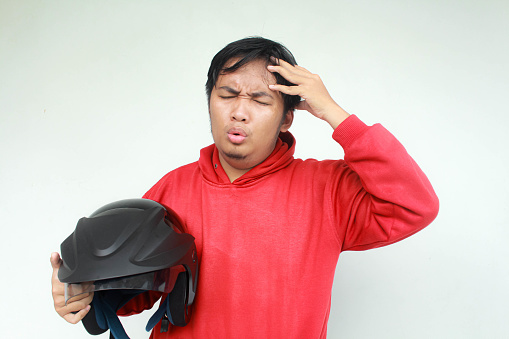 Young asian man holding his head with stressed and confused expression. While holding a motorcycle helmet. Safety concept.