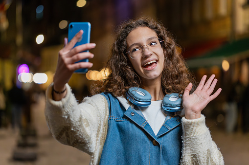 Happy teenage child girl blogger taking selfie on smartphone communicating video call online with subscribers recording stories for social media vlog. Young woman standing on city street at evening