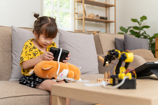 Happy small young girl sitting on a sofa using toy remote control to control arm robot to pick up and mode small wooden pieces around