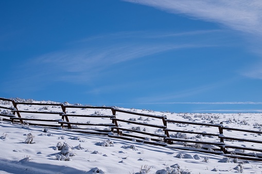 The image shows a snow-covered landscape with a zigzagging wooden fence, snow-dusted shrubbery, and a clear blue sky, creating a serene and tranquil winter scene.