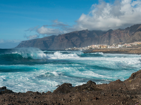 Big breaking waves splash against black lava rock coast. Turquoise blue Atlantic ocean with view of Los Gigantes cliffs and village. Tenerife, Canary islands, Spain. Sunny winter day