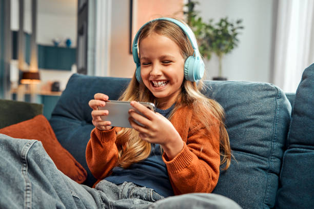 Entertainment in cyberspace. Laughing caucasian girl using mobile phone on sofa Entertainment in cyberspace. Laughing caucasian girl using bright headset and mobile phone while resting on sofa indoors. Careless preteen person watching comedy videos and having fun alone. free girl download stock pictures, royalty-free photos & images