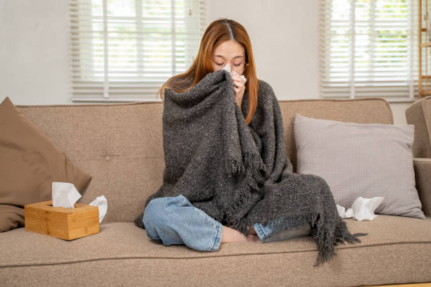 istock Young sick woman with soft blanket over her body blowing her nose into tissue paper while sitting cross legs on her sofa in her living room 2143576824