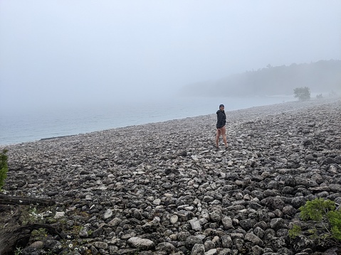 A wide shot of a rocky beach during a foggy day with a woman hiking
