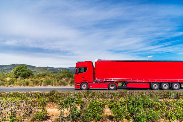 Truck with tauliner semi-trailer driving on a road between fields, completely red, side view.