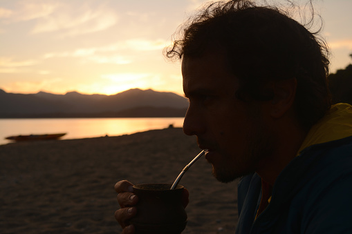 brown hair male person, drinking mate at sunset with lake in the background, vacation and relaxation