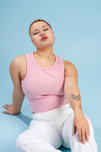 Woman wearing casual clothing with piercing, sitting on the floor in studio, closing eyes, isolated on blue background. Lgbt, body positive concept