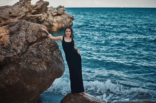 Woman in a black dress standing on a large rock on the seafront and leaning against another rock.