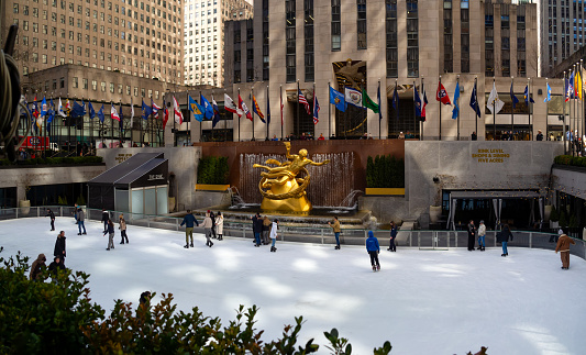 New York, USA - March 1, 2024: People enjoying Rockefeller Center Ice Skating Rink during winter, view of the iconic ice skating rink in New York open seasonally with state flags and Neptune statue.