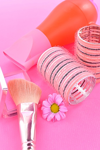 Beauty make-up routine essentials with a touch of floral grace on a vivid pink background and arrangement of beauty essentials on a pink backdrop, featuring a sleek hairdryer, sparkling hair rollers, a soft makeup brush, alongside a delicate pink flower and a blush palette. The composition radiates a playful yet chic vanity affair.