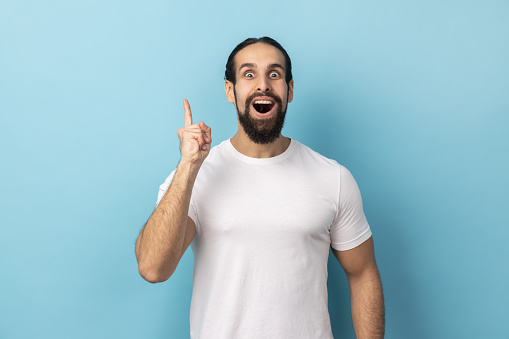 Portrait of excited amazed man with beard wearing white T-shirt gesturing finger up surprised by genius idea, got sudden clever solution. Indoor studio shot isolated on blue background.