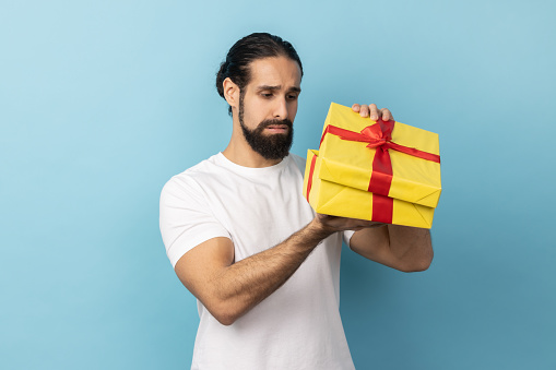 Portrait of unhappy man with beard wearing white T-shirt looking inside present box and expressing sadness, hoped to get another gift. Indoor studio shot isolated on blue background.