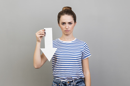 Portrait of sad upset woman wearing striped T-shirt showing white arrow pointing down, expressing sadness and sorrow, downgrade concept. Indoor studio shot isolated on gray background.