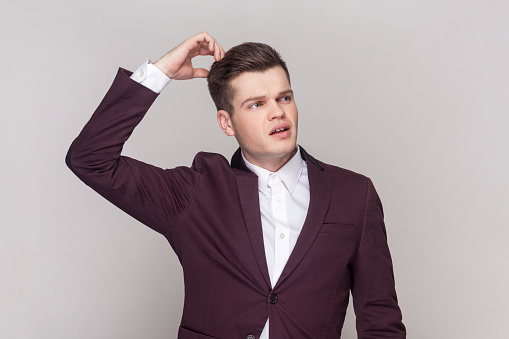 Portrait of contemplative handsome young man rubbing head as thinks, makes decision, focused away, wearing violet suit and white shirt. Indoor studio shot isolated on grey background.