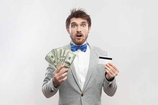 Portrait of shocked surprised attractive bearded man standing holding dollar banknotes and credit card, wearing grey suit and blue bow tie. Indoor studio shot isolated on gray background.