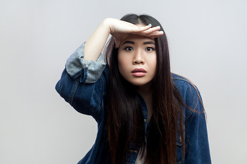 Portrait of woman in blue denim jacket standing keeping palm over head and looking attentively far away, peering with expectation at long distance. Indoor studio shot isolated on gray background.