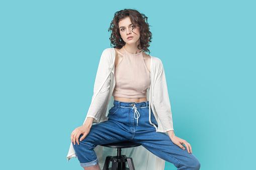 Portrait of confident beautiful dark haired woman sitting on chair and posing like fashion model and looking at camera with serious face. Indoor studio shot isolated on blue background.
