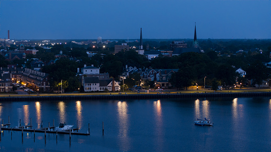 Illuminated coastal neighborhood at night. Mix of wealthy townhouses, parking lots, and residential houses on the riverbank. Downtown Portsmouth, Virginia in the distant. Aerial view