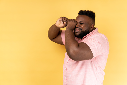 Portrait of handsome bearded man wearing pink shirt making glasses shape, looking through binoculars gesture with attentive expression. Indoor studio shot isolated on yellow background.