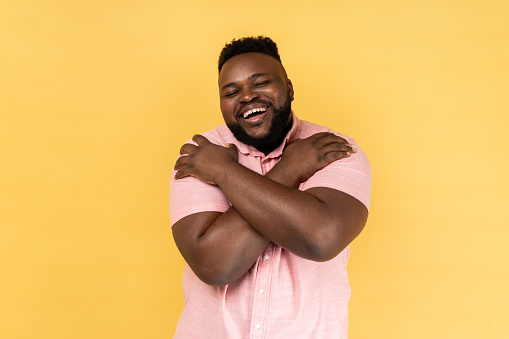 Portrait of selfish narcissistic man wearing pink shirt embracing himself and smiling with expression of great ego, pleasure and self-esteem. Indoor studio shot isolated on yellow background.