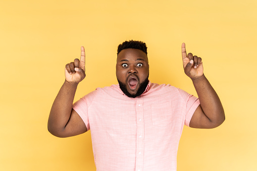 Wow, attention to advertising above! Portrait of surprised man wearing pink shirt pointing up at advertisement area, copy space for promotional text. Indoor studio shot isolated on yellow background.
