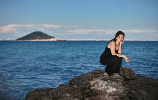 Sentimental woman in a black dress sitting on a rock on the beach at sunset.