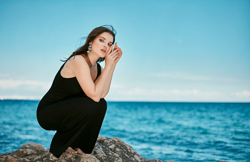 Sentimental woman in a black dress sitting on a rock on the beach at sunset.