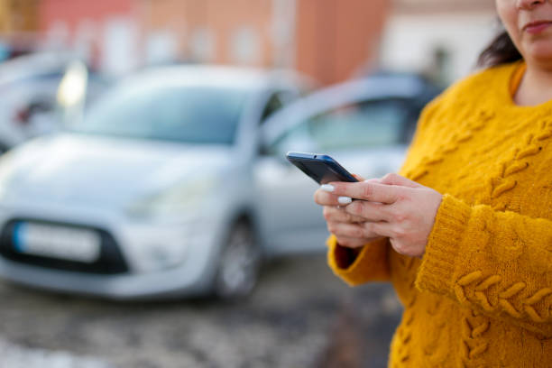 istock A woman is holding a cell phone while standing next to a silver car. She is focused on her phone, possibly texting or browsing the internet 2144560175