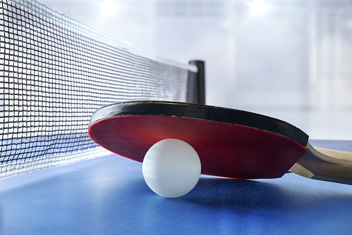 Ping pong paddle resting on white ball on a blue game table next to the game net with sports pavilion with lighting in the background. Front view.