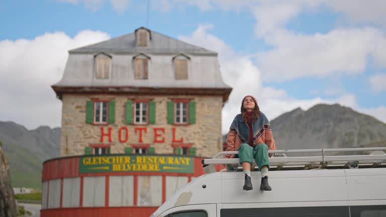 Woman sitting on camper van roof  near the old hotel on Furka Pass in Alps