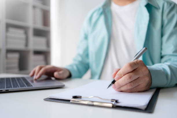 istock A professional is engaged in writing notes on a clipboard while using a laptop in a modern office setting. 2144885815