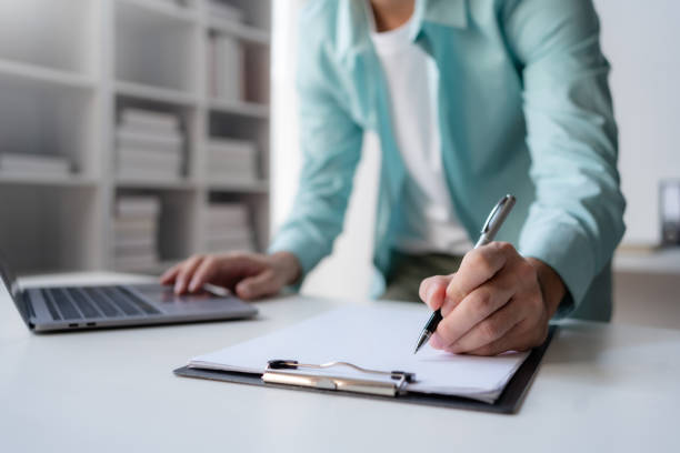 istock A professional is engaged in writing notes on a clipboard while using a laptop in a modern office setting. 2144885819