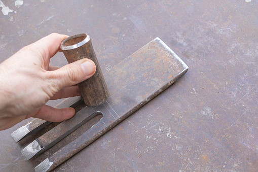 A hand holds a round steel bar with straight edges and a flat top, next to it is an iron chisel.