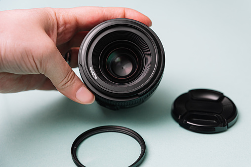 Close-up of a hand holding a DSLR lens with its cap and a UV filter placed beside it on a soft teal background.