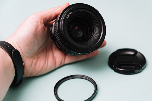 A person's hand displaying a DSLR camera lens with a UV filter and lens cap set aside, against a soft turquoise background.