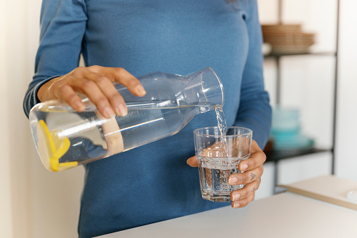 Young beautiful African American woman pouring clean water into glass in kitchen. Attractive active thirsty girl drink or take a sips of mineral water for health care, wellbeing after sport