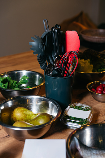 Assorted cooking utensils in a container with bowls of fresh ingredients ready for meal preparation on a wooden table.