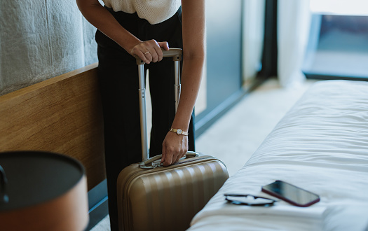 Anonymous woman standing in her hotel room zipping up her suitcase ready to leave. There is a mobile phone and sunglasses on the bed next to her.