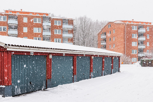 A row of red and blue residential buildings in Mölndal, Sweden, covered in a blanket of snow. In the foreground, there is a car garage with snowfall gently drifting down.