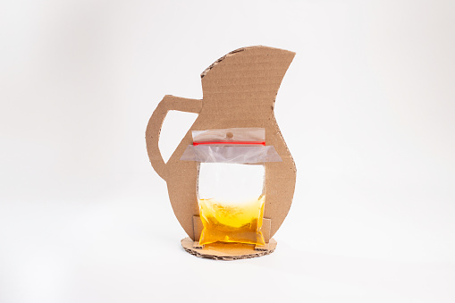 Cardboard pitcher craft: plastic bag with yellow colored water affixed to cut out piece, backside view, white background