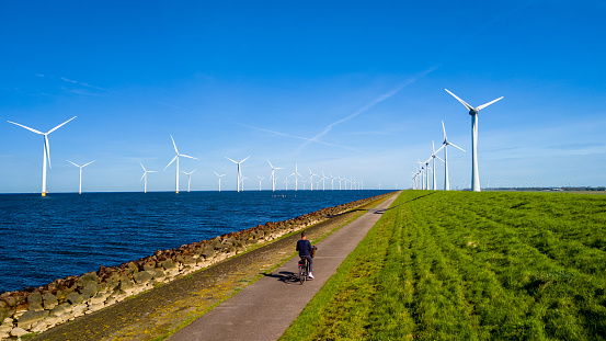 A person gracefully pedals a bike down a path alongside a calm body of water, with windmill turbines in the distance in the Netherlands, men on green electric bike
