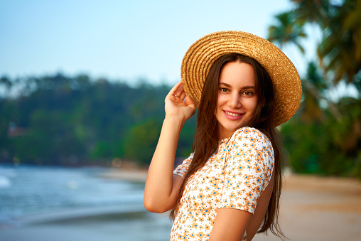 Summer fashion, leisure by sea, female traveler explores coast, poses with ocean backdrop, retro vibe in holiday attire. Smiling woman in floral dress and straw hat enjoys tropical beach at sunset.