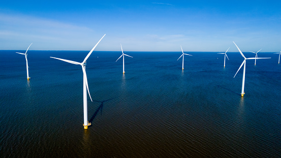 A group of wind turbines elegantly spin in the ocean off the coast of the Netherlands in Flevoland during a tranquil spring day. drone aerial view of windmill turbines green energy in the ocean