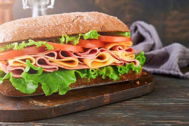 istock Sandwich. One fresh big submarine sandwich with ham, cheese, lettuce, tomatoes and microgreens on old wooden dark background. Healthy breakfast theme concept, school lunch, breakfast or snack. 2145276771
