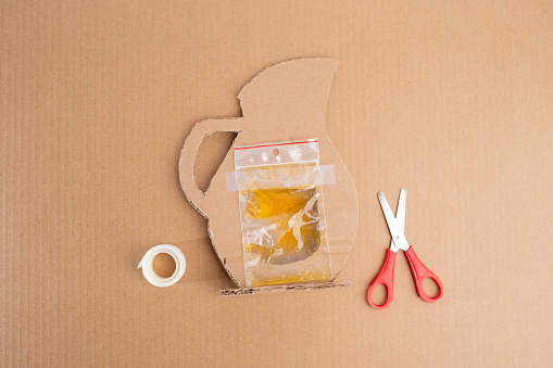 Rear side perspective of inventive cardboard crafting: plastic bag containing yellow liquid affixed to cardboard pitcher, top view