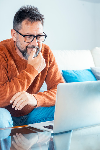 One adult freelance man looking the laptop screen thinking and touching chin. Wearing glasses mature male. Home work smart working remote job business concept lifestyle. Modern worker sitting on sofa
